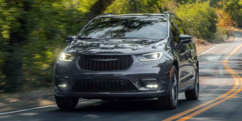 The image showcases a sleek gray Chrysler Pacifica, driving on a scenic road surrounded by lush greenery, highlighting its modern and stylish design, perfect for family journeys.

