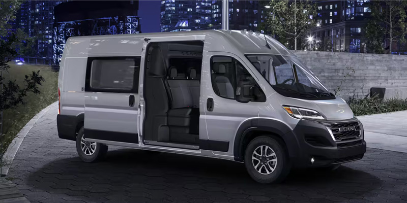 The image showcases a silver Ram ProMaster van, parked in an urban setting with the sliding door open, highlighting its spacious interior and versatile design, ideal for both work and leisure purposes.
