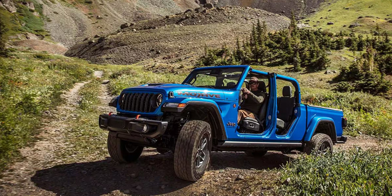 Blue Jeep Gladiator pickup driving off-road on a rocky mountain trail with doors removed.
