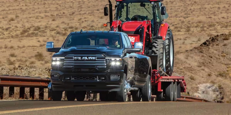 2026 Ram 3500 heavy-duty pickup truck towing a red tractor on a flatbed trailer through a rugged desert landscape.
